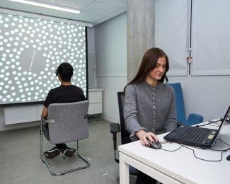 A female student works at a laptop at a desk while a person behind her is facing a screen covered in dots as part of an experiment.